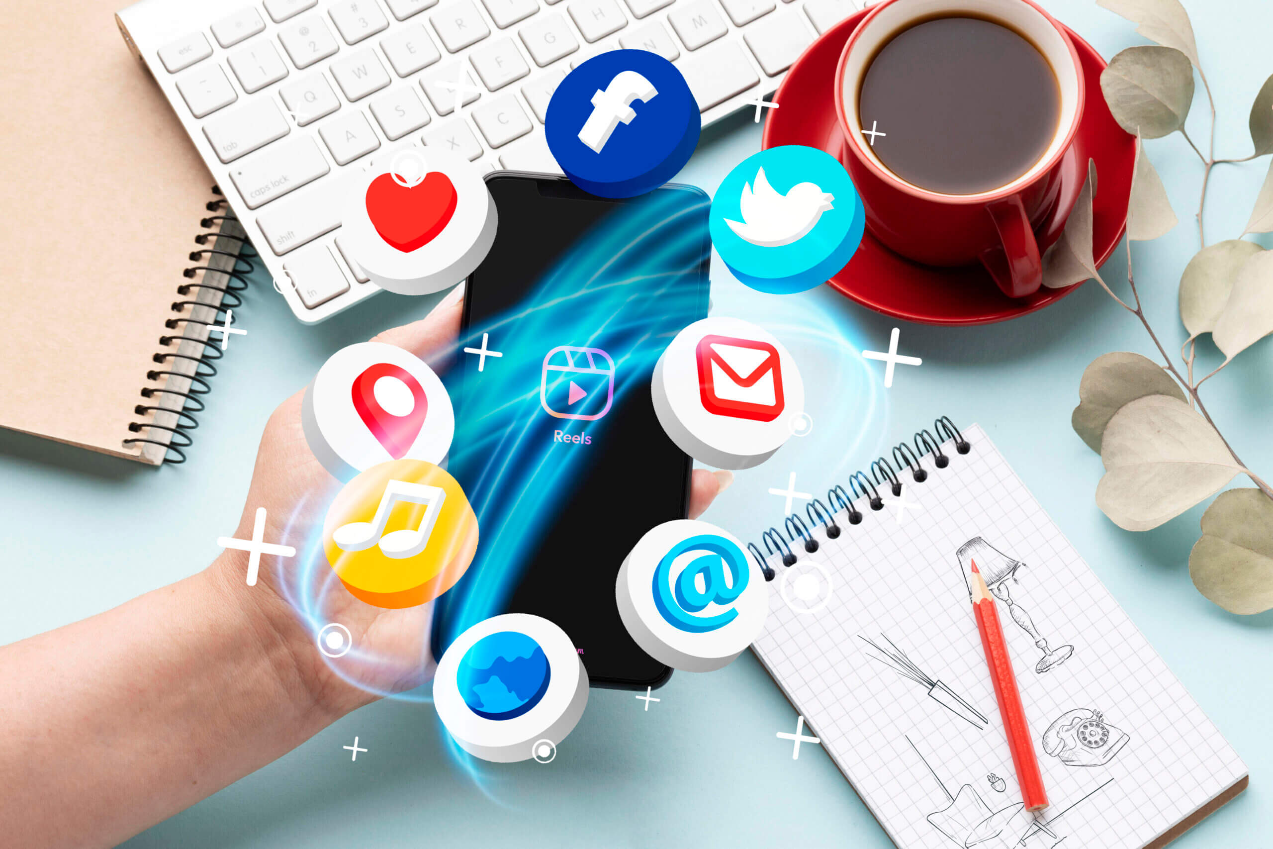 A hand holding a smartphone with floating social media icons, including Facebook and Twitter, surrounded by a keyboard, notepad, coffee cup, and plant.